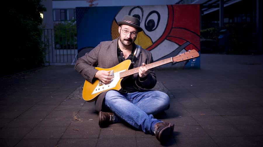 Mann sitzt mit Gitarre und Hut auf dem Boden im Freien